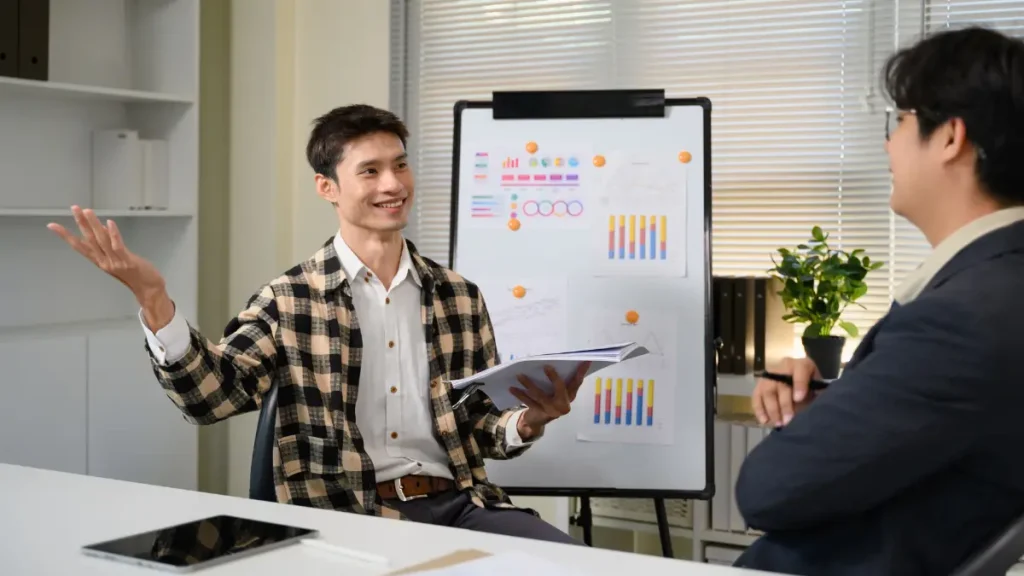 Seorang pengusaha muda sedang melakukan pitching ide dalam bahasa Inggris dengan penuh percaya diri di depan para eksekutif di kantor modern.